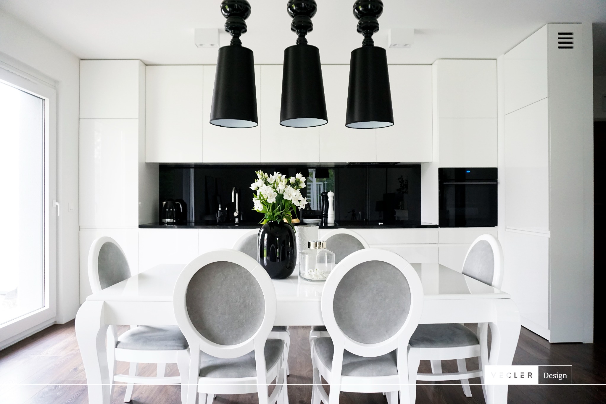 Biała, minimalistyczna jadalnia z okrągłymi krzesłami i czarnymi lampami wiszącymi, kwiaty w wazonie na stole, czarny panel nad blatem kuchennym.