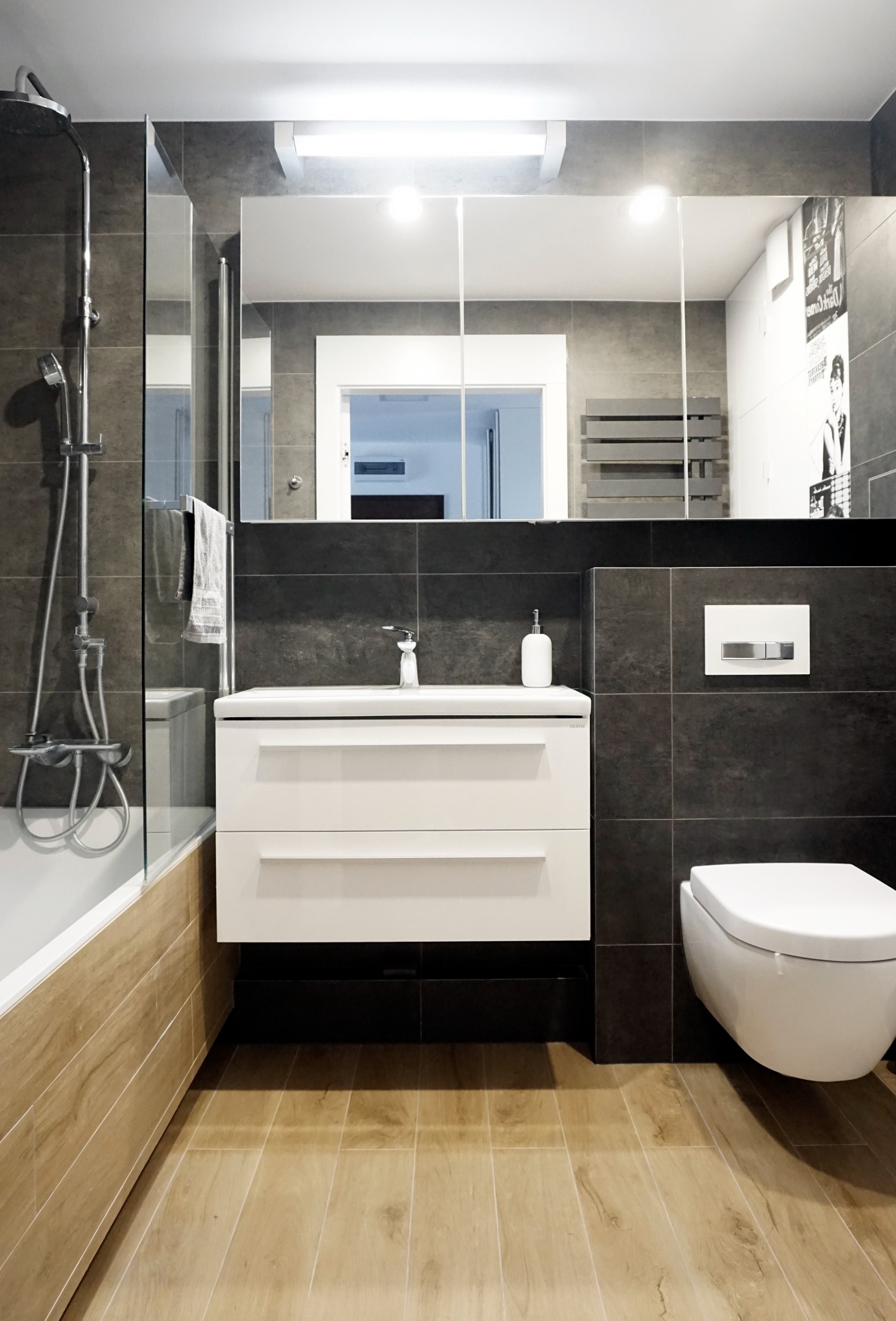 Nowoczesna łazienka z ciemnymi płytkami, białymi meblami i drewnopodobną podłogą, widoczna bateria umywalkowa z wysoką wylewką i wisząca toaleta