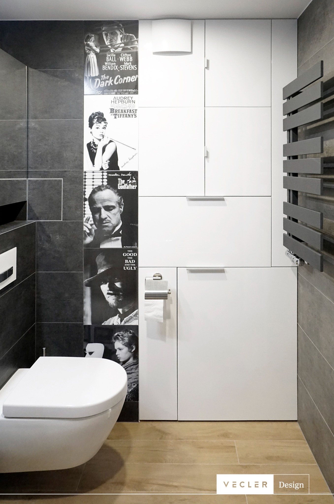 Nowoczesna łazienka z wiszącą toaletą, grzejnikiem drabinkowym i szafkami w zabudowie. Ściana zdobiona czarno-białymi plakatami filmowymi w stylu vintage.