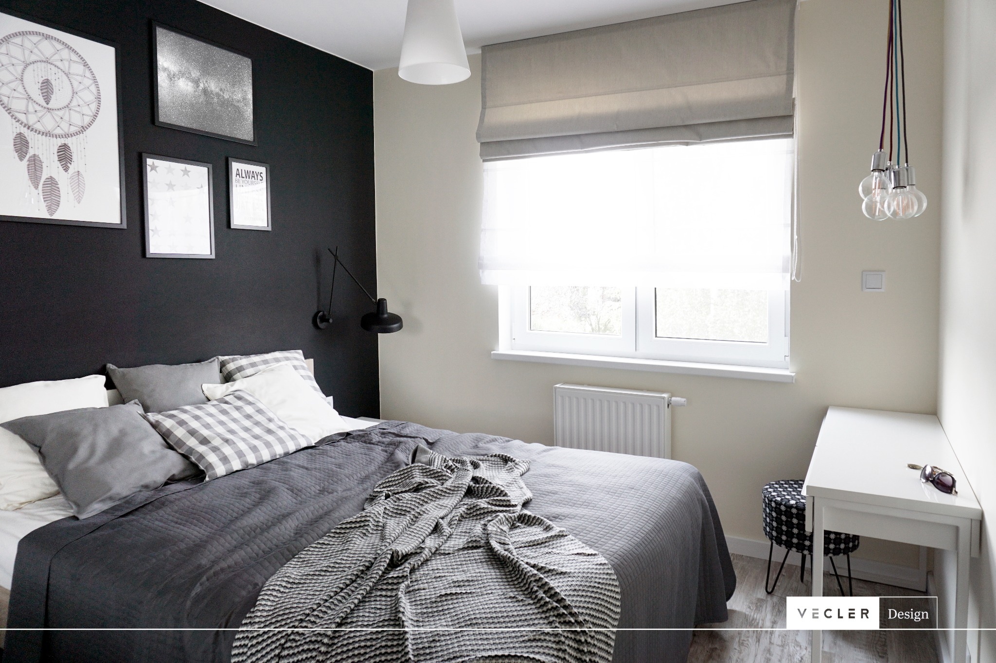 Nowoczesna sypialnia z czarną ścianą, galerią obrazów, szarą pościelą i dekoracyjnym oświetleniem, uzupełniona o białe biurko i wzorzysty stołek.