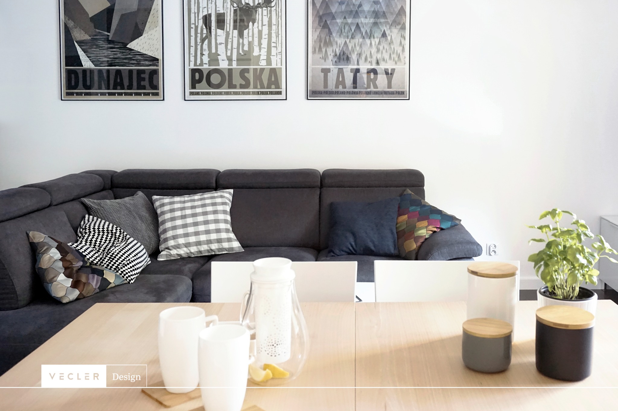 Szary narożnik z poduszkami w salonie, nad nim trzy plakaty z motywami krajobrazowymi Polski: Dunajec, Polska, Tatry. Na drewnianym stole białe kubki, dzbanek z cytryną i dekoracyjne pojemniki...
