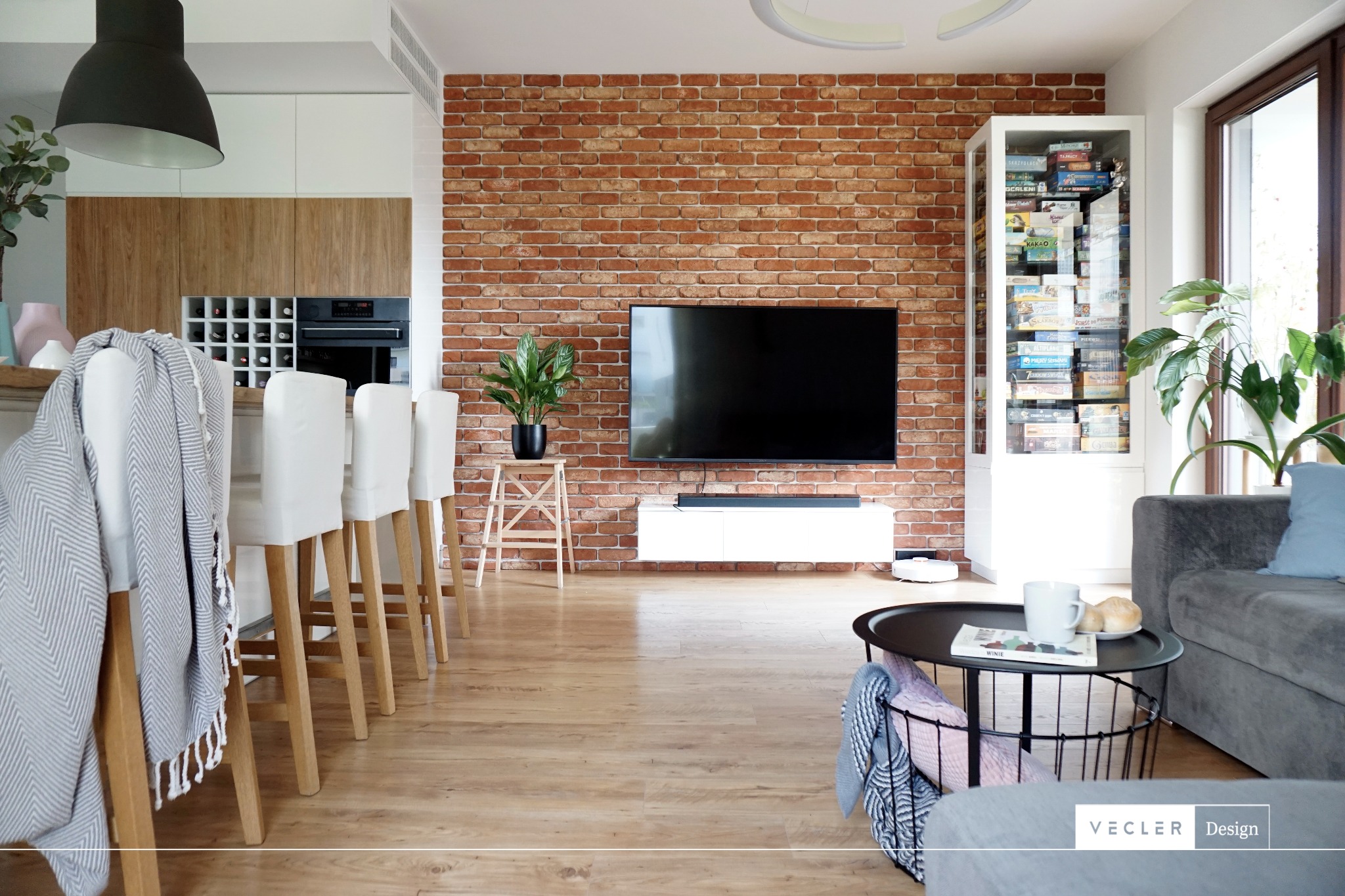 Salon z ceglaną ścianą, telewizorem, szafką RTV, regałem z grami planszowymi, szarym narożnikiem i stolikiem kawowym z filiżanką oraz krzesłami barowymi z białymi pokrowcami i szarym pledem.