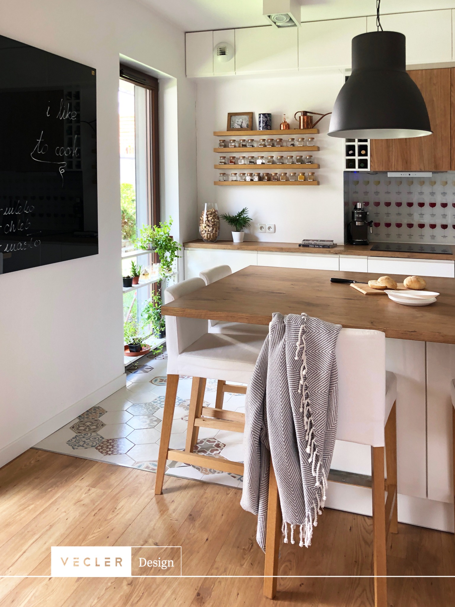 Jasna kuchnia w stylu skandynawskim z drewnianym blatem wyspy, dekoracyjnymi płytkami podłogowymi i czarną tablicą z napisem 'I like to cook'.