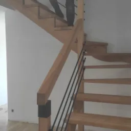 Jasne, drewniane schody z czarnymi, metalowymi balustradami w nowoczesnym domu. Osoba wchodzi po schodach, trzymając w ręku tablet.