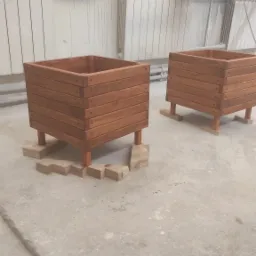 Dwie drewniane, lakierowane donice w kształcie sześcianu na nóżkach, stojące na betonowej posadzce w warsztacie, podparte drewnianymi klockami.