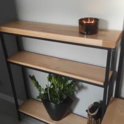 Trzypoziomowy regał z jasnego drewna i czarnych metalowych elementów, dekorowany świecą zapachową, rośliną w czarnej doniczce i lampionem, ustawiony na szarym panelu podłogowym, z fragmentem...