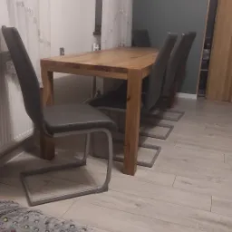 Drewniany stół jadalny z czterema krzesłami z szarą tapicerką i metalowymi nogami, ustawiony na podłodze z paneli, z widocznym fragmentem dywanu w rogu kadru.