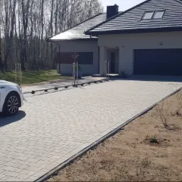 Nowoczesny dom z szarą kostką brukową na podjeździe, biały samochód zaparkowany obok, młode drzewka posadzone wzdłuż podjazdu, las w tle.