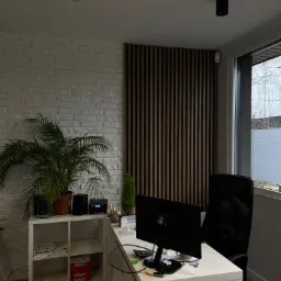 Nowoczesne biuro domowe z białymi cegłami na ścianie, drewnianymi panelami ściennymi, biurkiem z komputerem i roślinami doniczkowymi.