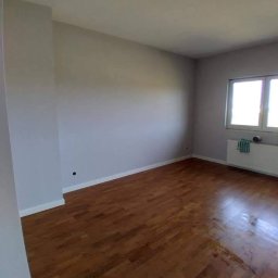 HouseGello Aleh Kapranau - Pokój po malowaniu ścian na szaro, z drewnianą podłogą i grzejnikiem pod oknem, na którym wisi ręcznik.