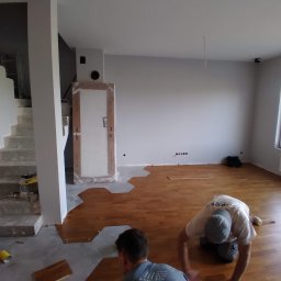 HouseGello Aleh Kapranau - Układanie parkietu w jasnym pomieszczeniu z widocznymi schodami i zapakowanymi drzwiami, dwóch mężczyzn w trakcie pracy z drewnianymi klepkami i gumowym młotkiem.