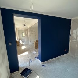 HouseGello Aleh Kapranau - Świeżo pomalowany na granatowo pokój z widoczną kuwetą malarską na podłodze i otwartym przejściem do kolejnego pomieszczenia w trakcie prac wykończeniowych.