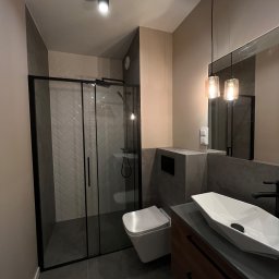 HouseGello Aleh Kapranau - Nowoczesna łazienka z kabiną prysznicową z czarnymi ramami, białą armaturą, umywalką nablatową o geometrycznym kształcie i wiszącą toaletą, oświetlona dwoma wiszącymi lampami z bursztynowego szkła.