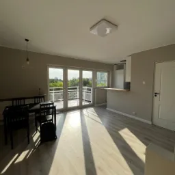 Jasne, przestronne wnętrze mieszkania z parkietem w odcieniach beżu, czarnym stołem z krzesłami, dużymi oknami balkonowymi wpuszczającymi silne promienie słoneczne i widokiem na zieleń,...