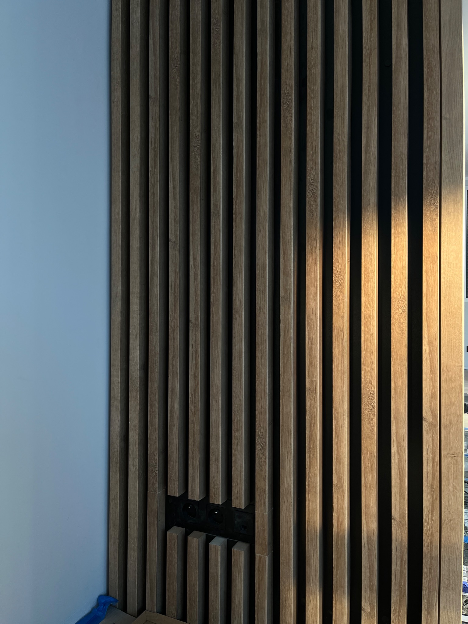 Drewniane lamele ścienne w kolorze jasnego dębu, zamontowane na ścianie obok gładkiej, błękitnej powierzchni, z widocznym gniazdkiem elektrycznym wkomponowanym w strukturę lameli.