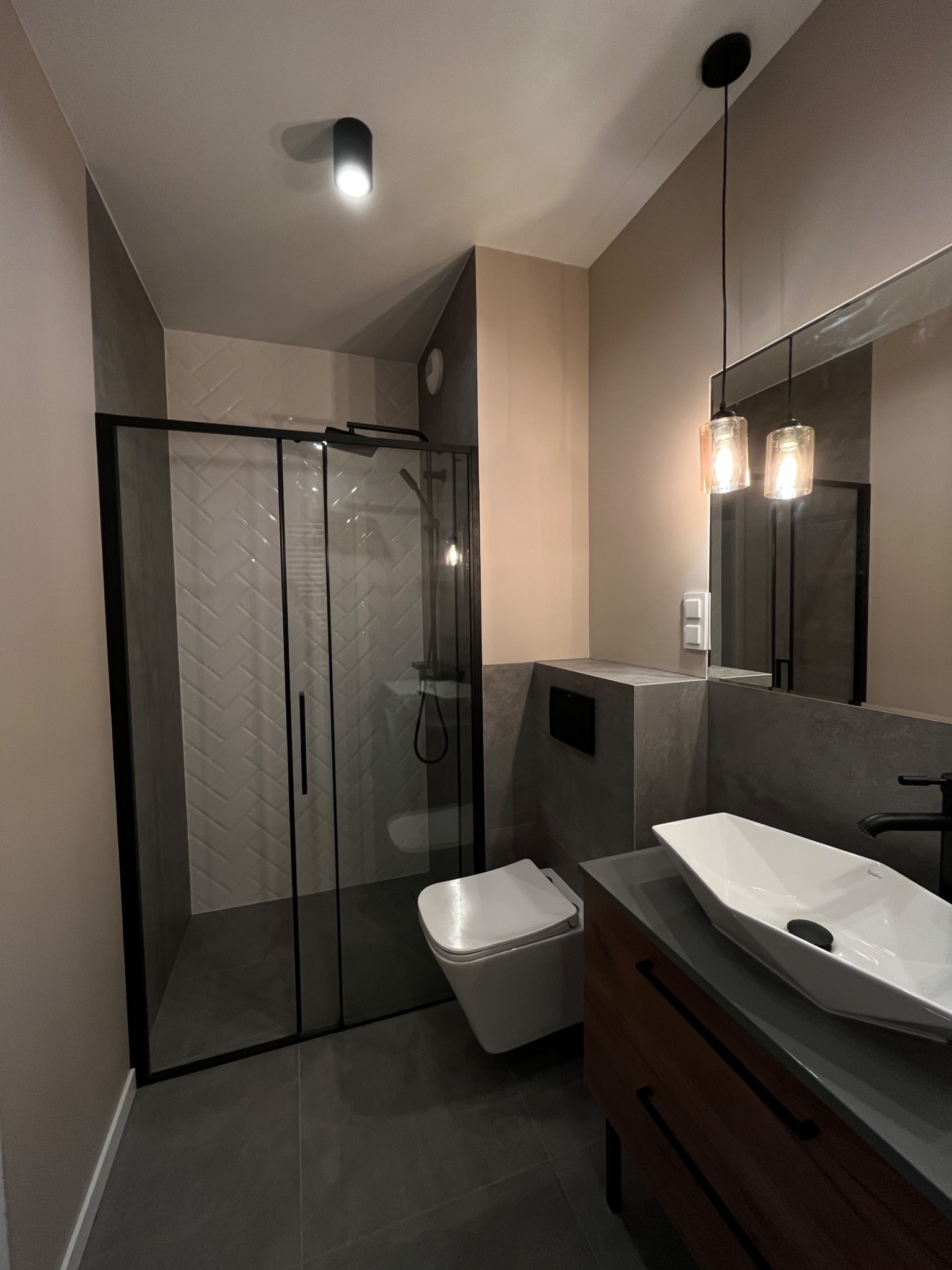 Nowoczesna łazienka z kabiną prysznicową z czarnymi ramami, białą armaturą, umywalką nablatową o geometrycznym kształcie i wiszącą toaletą, oświetlona dwoma wiszącymi lampami z bursztynowego szkła.