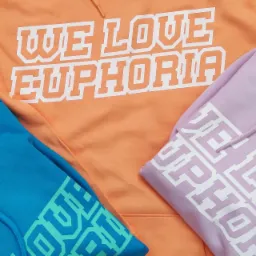 Trzy kolorowe bluzy z kapturem: pomarańczowa z napisem 'WE LOVE EUPHORIA', liliowa i turkusowa z nadrukami na rękawach, ułożone na płasko.