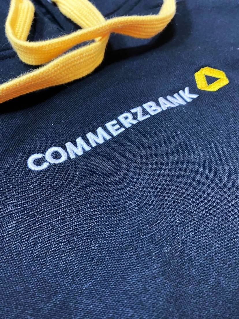 Granatowa bluza z kapturem, z wyhaftowanym białym napisem Commerzbank i żółtym logo, oraz żółtym sznurkiem kaptura.