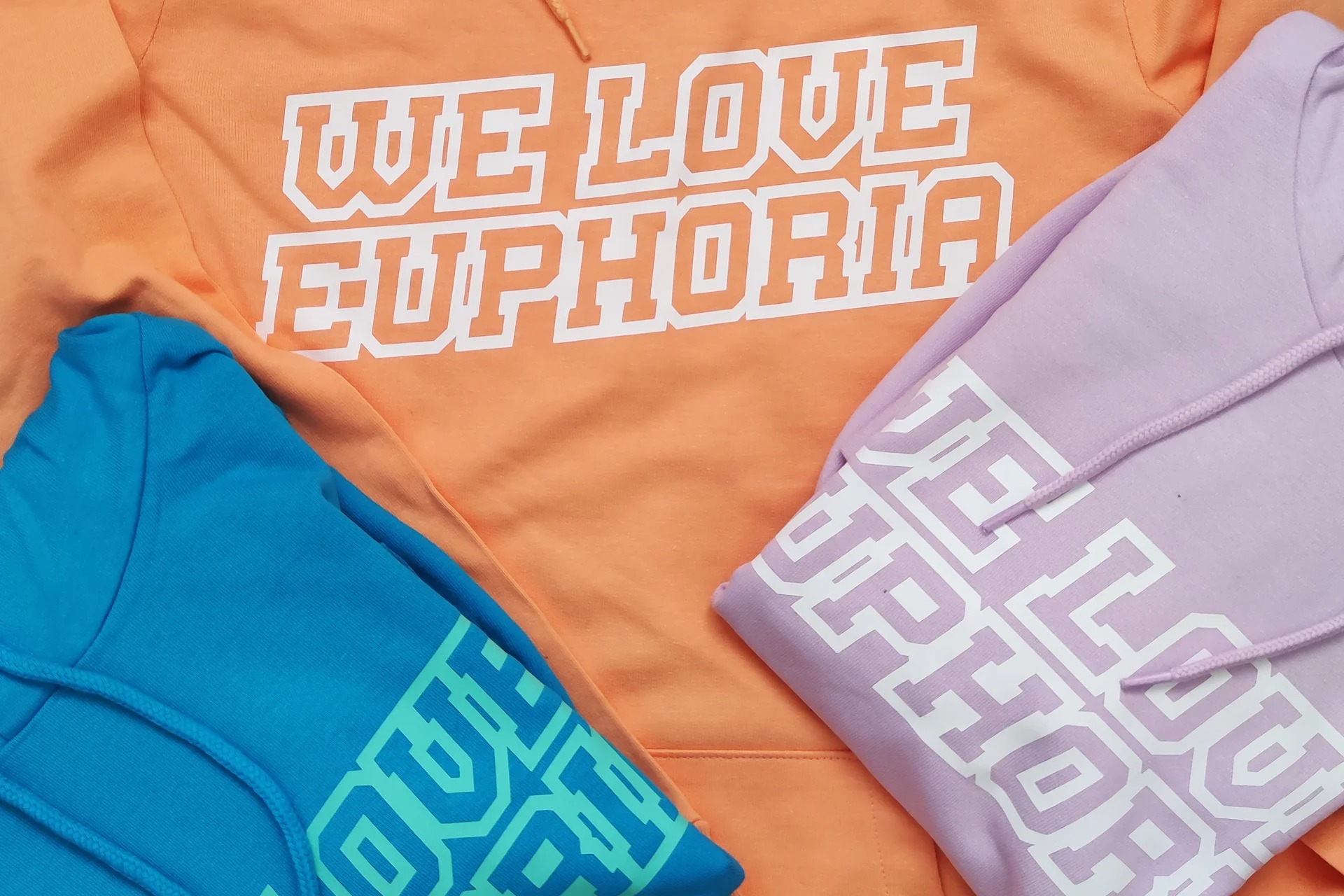 Trzy kolorowe bluzy z kapturem: pomarańczowa z napisem 'WE LOVE EUPHORIA', liliowa i turkusowa z nadrukami na rękawach, ułożone na płasko.