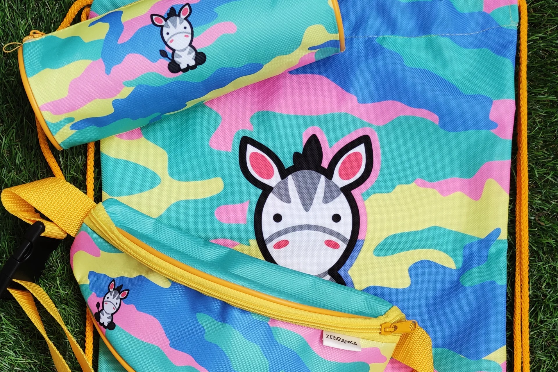 Zestaw: plecak-worek, piórnik-tubka i saszetka biodrowa z kolorowym wzorem kamuflażu i motywem zebry na tle zielonej trawy.