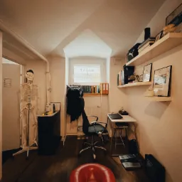 Gabinet fizjoterapeuty: czerwony zagłówek do masażu na pierwszym planie, w tle szkielet, biurko z laptopem i półki z książkami, oświetlenie górne.