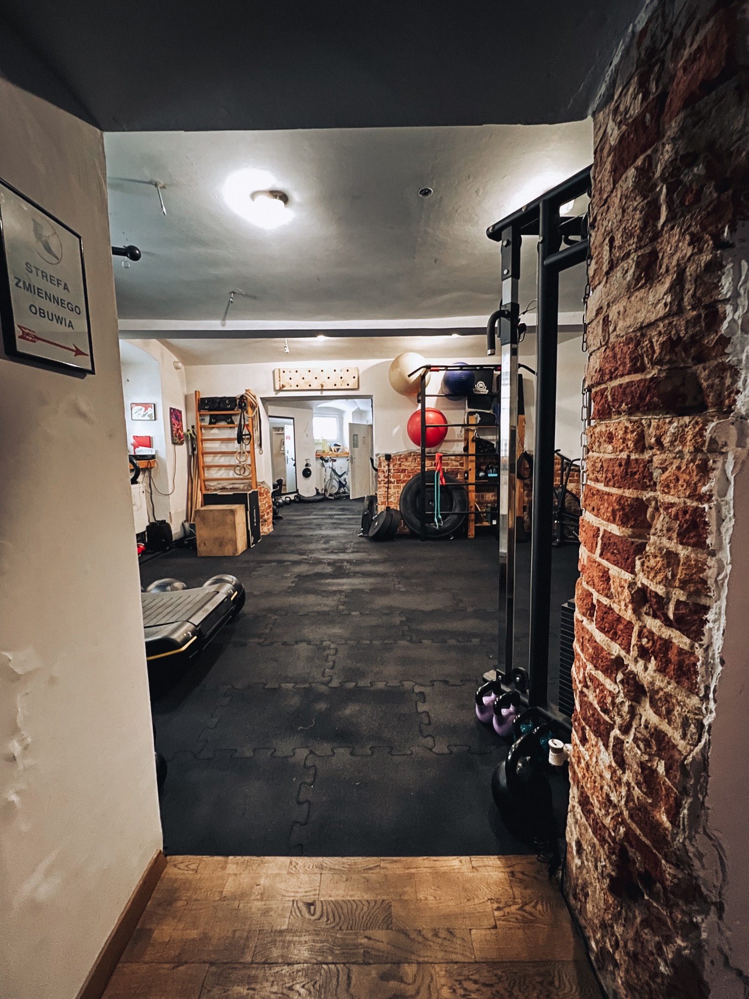 Widok na salę ćwiczeń z różnorodnym sprzętem, w tle drabinki gimnastyczne i akcesoria, z boku kolumna z czerwonej cegły.