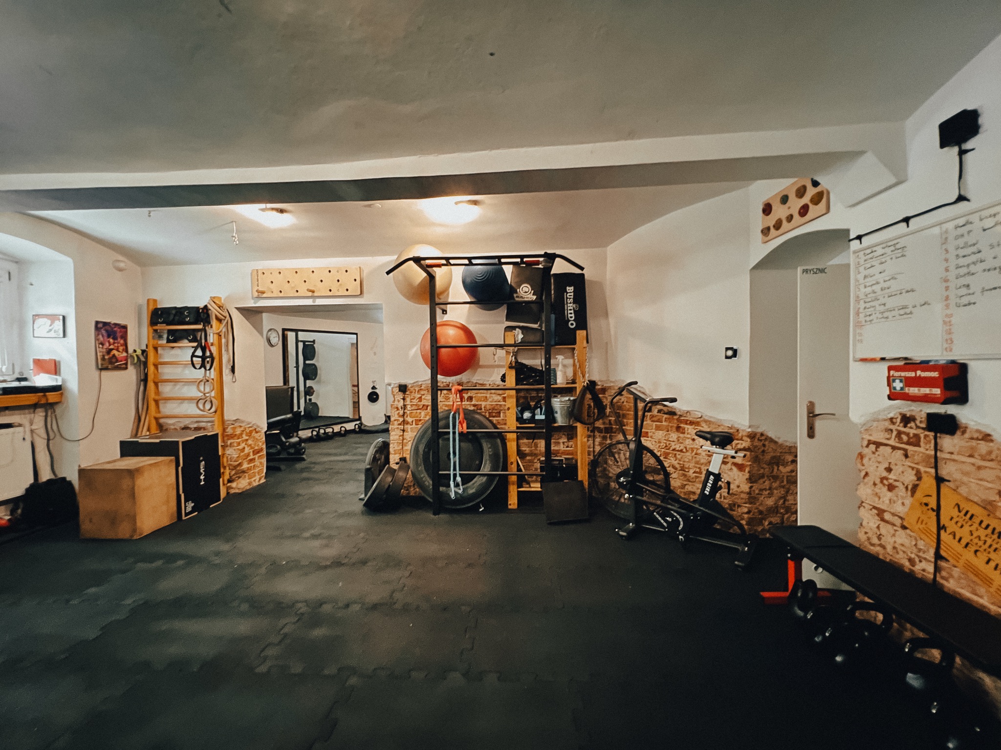 Domowa siłownia z wyposażeniem: drabinka gimnastyczna, sztanga, ciężary, rower treningowy, piłki do ćwiczeń i tablica z planem treningowym na tle ściany z cegły.