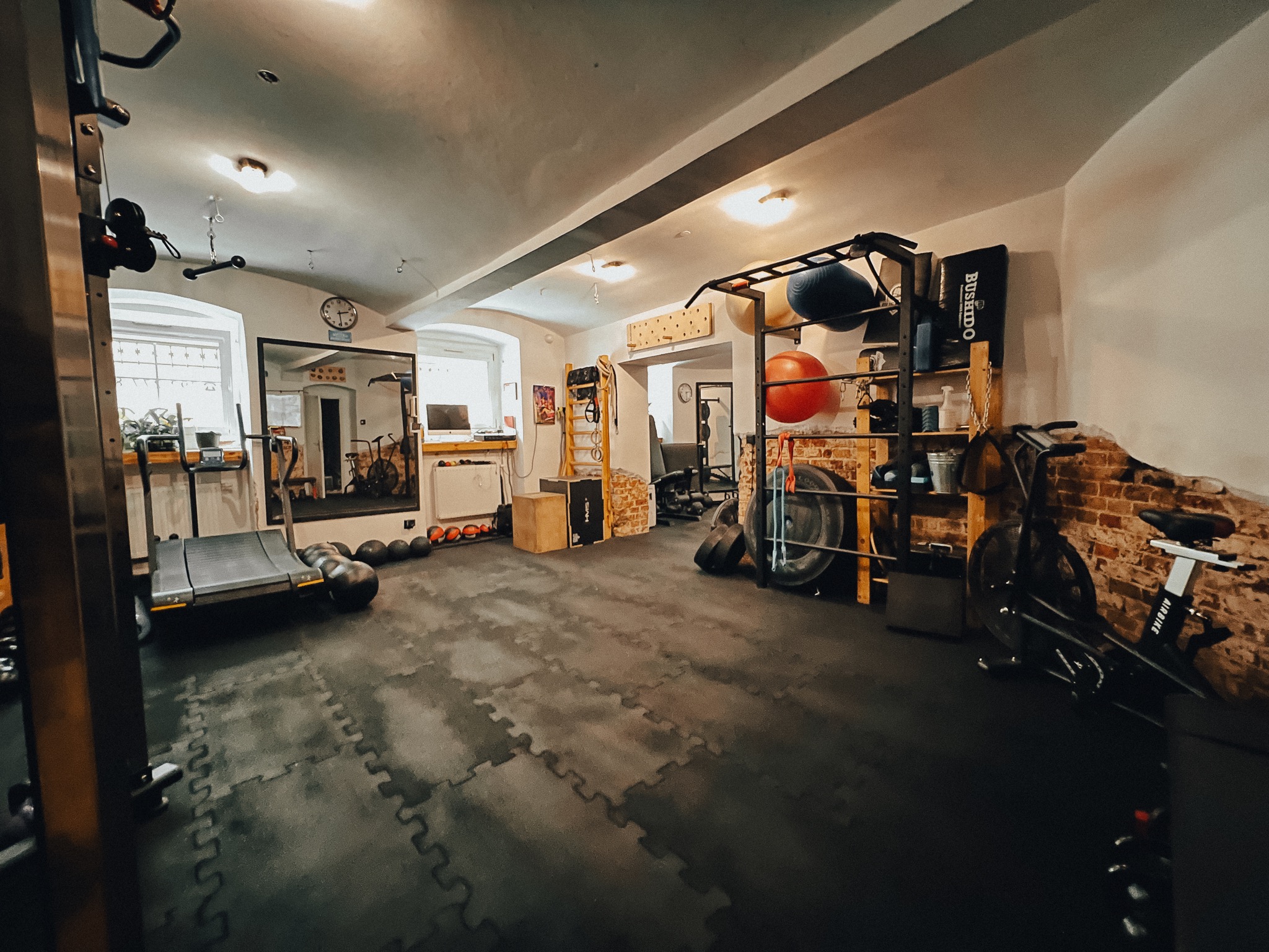 Przestronna, dobrze wyposażona siłownia domowa z bieżnią, rowerem stacjonarnym Airbike, piłkami gimnastycznymi, drabinką gimnastyczną i podłogą z mat piankowych, oświetlona lampami sufitowymi.