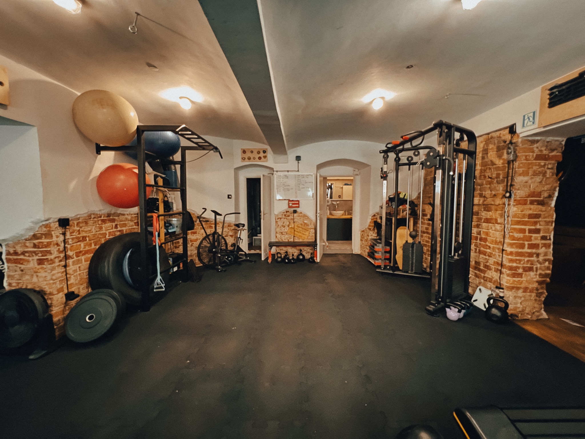 Przestronna siłownia w piwnicy z ceglanymi ścianami, wyposażona w sprzęt do ćwiczeń, w tym maszyny siłowe, piłki, ciężary i rowerek treningowy.