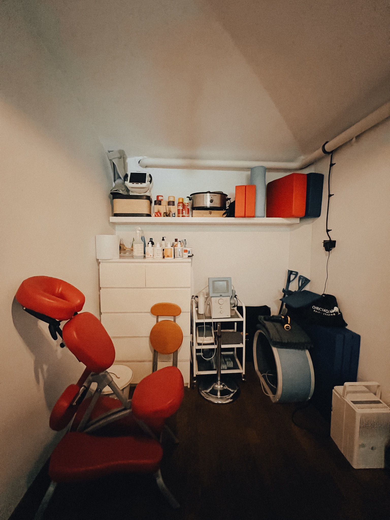 Wnętrze gabinetu z czerwonym krzesłem do masażu na pierwszym planie, widoczny regał z aparaturą medyczną, biała komoda z kosmetykami i półka z akcesoriami do ćwiczeń.