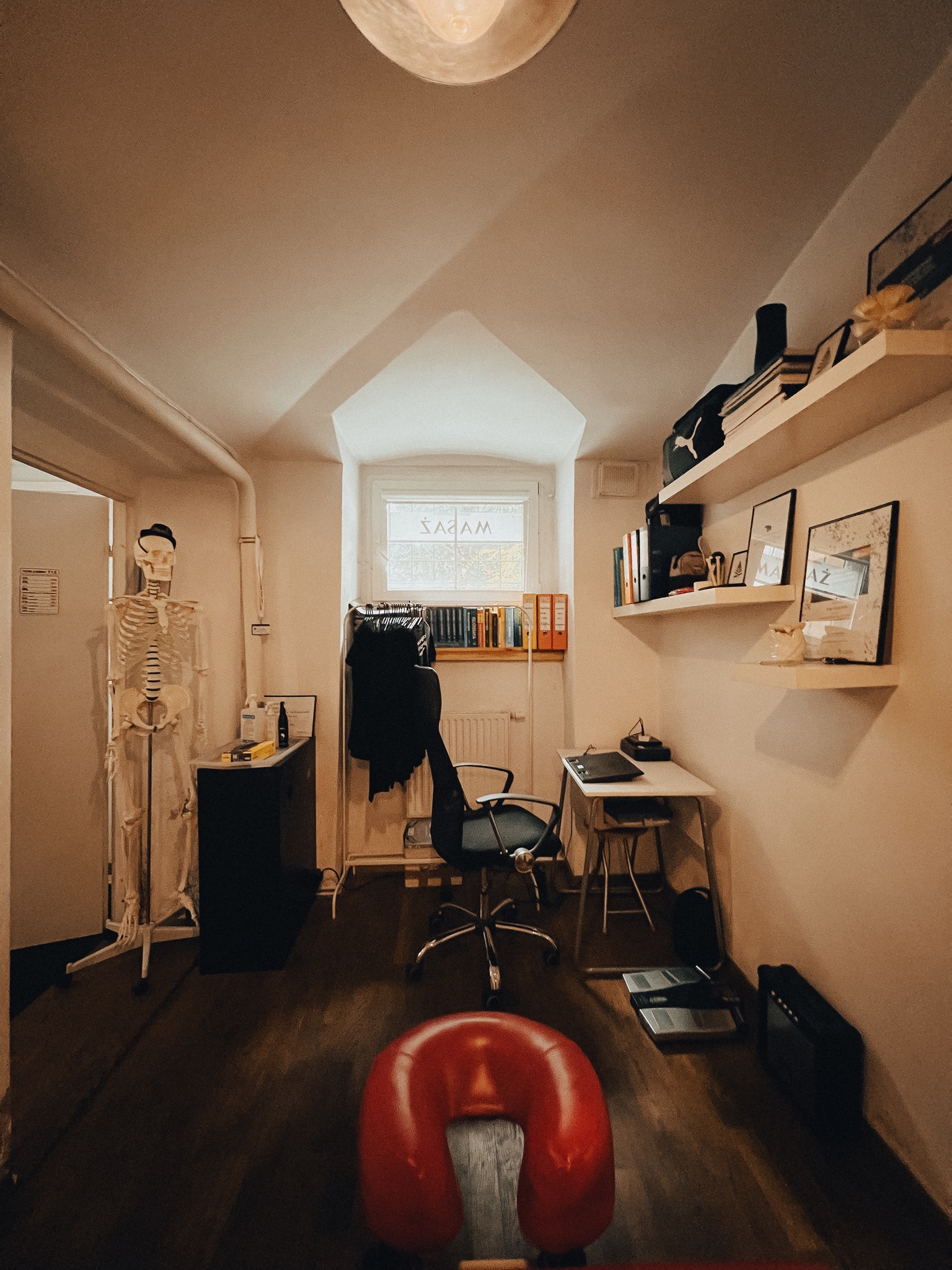 Gabinet fizjoterapeuty: czerwony zagłówek do masażu na pierwszym planie, w tle szkielet, biurko z laptopem i półki z książkami, oświetlenie górne.