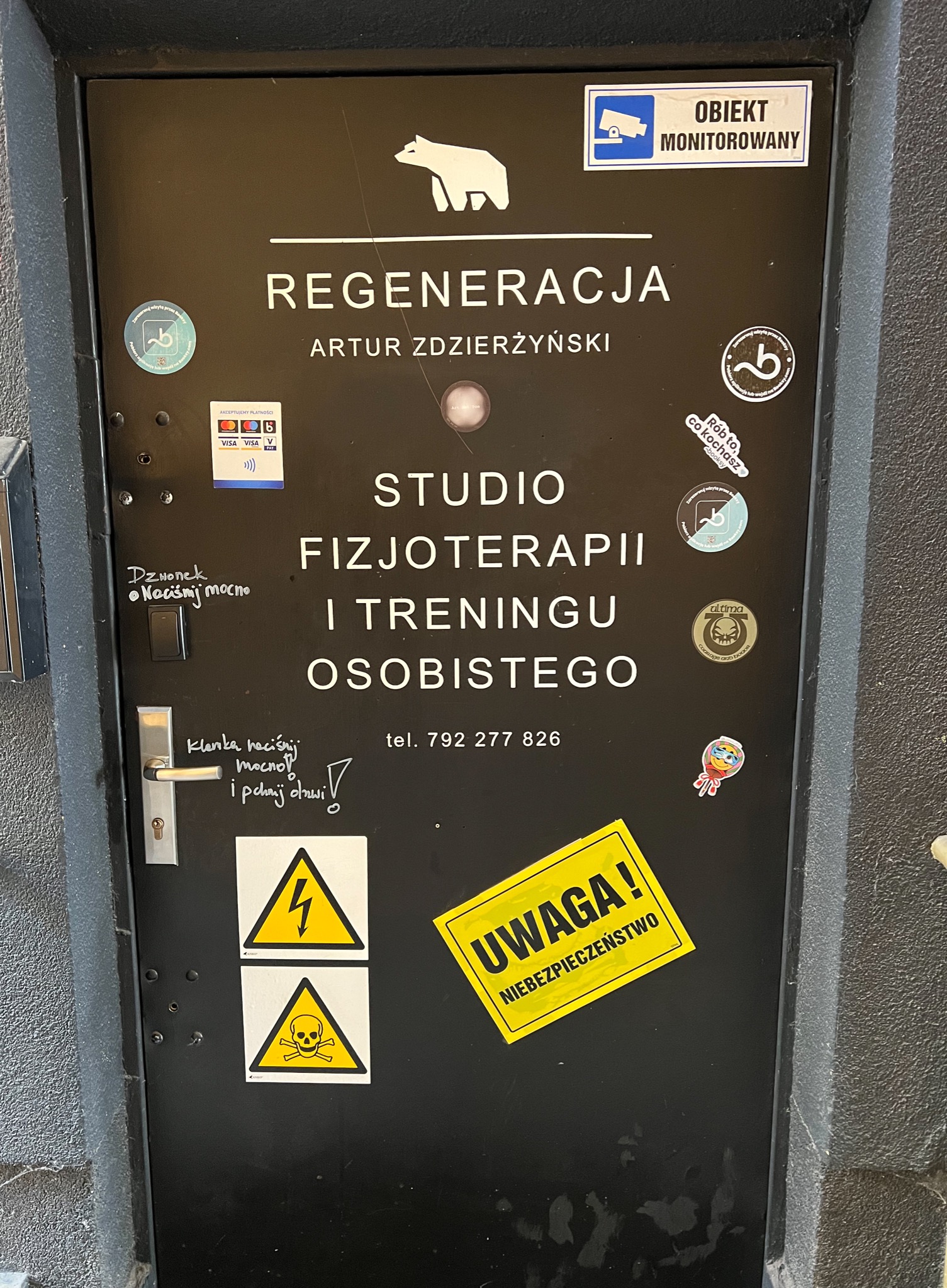 Czarne drzwi wejściowe do studio 'Regeneracja Artur Zdzierzyński' oferującego fizjoterapię i trening osobisty, z naklejkami informacyjnymi, ostrzegawczymi i logo, oraz odręcznymi notatkami...