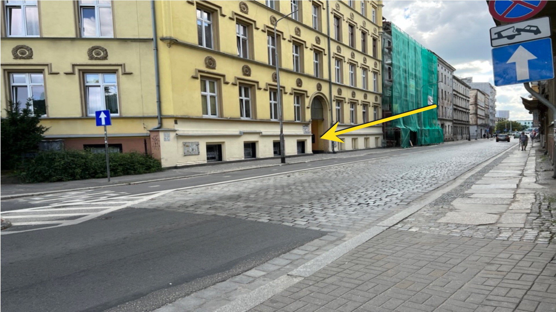 Ulica z żółtą kamienicą w tle, chodnik wyłożony kostką, budynek w remoncie pod zieloną siatką, znak drogowy z niebieską strzałką.