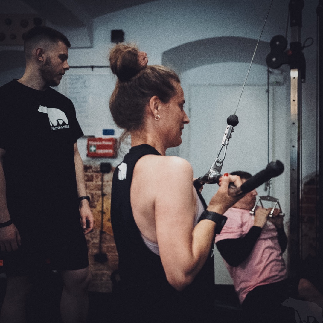 Kobieta ćwiczy na maszynie wyciąg górny pod okiem mężczyzny w czarnej koszulce z białym niedźwiedziem, w tle ćwiczy druga osoba.