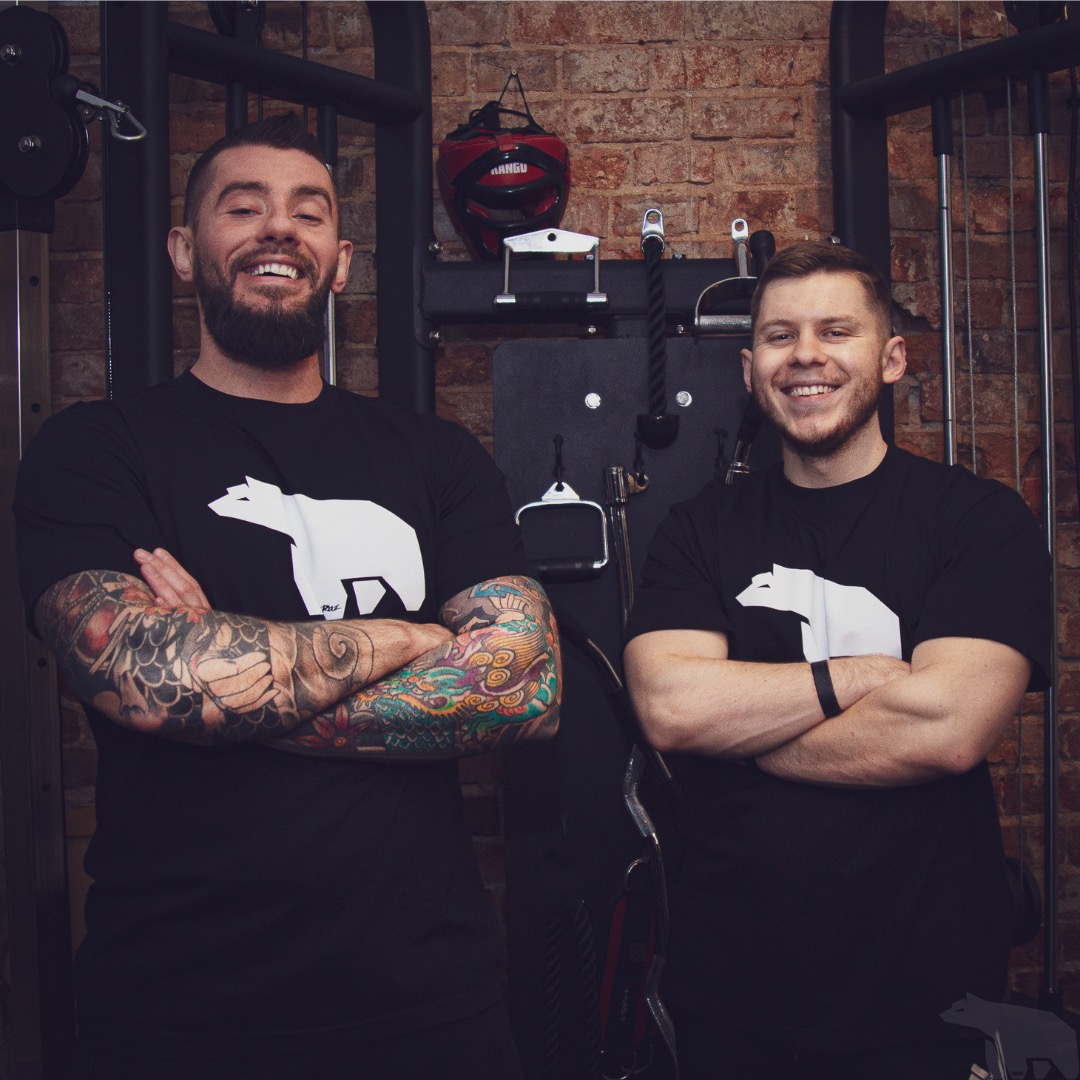 Dwóch uśmiechniętych trenerów personalnych w czarnych koszulkach z logo białego niedźwiedzia, stojących na tle ceglanej ściany i sprzętu do ćwiczeń.