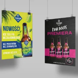 Dwa plakaty reklamowe wiszące na sznurkach za pomocą klipsów. Jeden z plakatem piwa bezalkoholowego o smaku ice tea, drugi z plakatem piwa Pink Boots. Oba plakaty mają ciemne tło u góry...