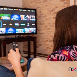 Oxylion Sp. z o.o. - Kobieta z tyłu siedzi na kanapie, trzyma pilot i ogląda telewizor z interfejsem platformy streamingowej na tle ceglanej ściany.