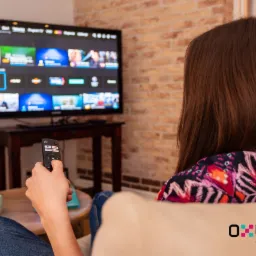 Kobieta z tyłu siedzi na kanapie, trzyma pilot i ogląda telewizor z interfejsem platformy streamingowej na tle ceglanej ściany.