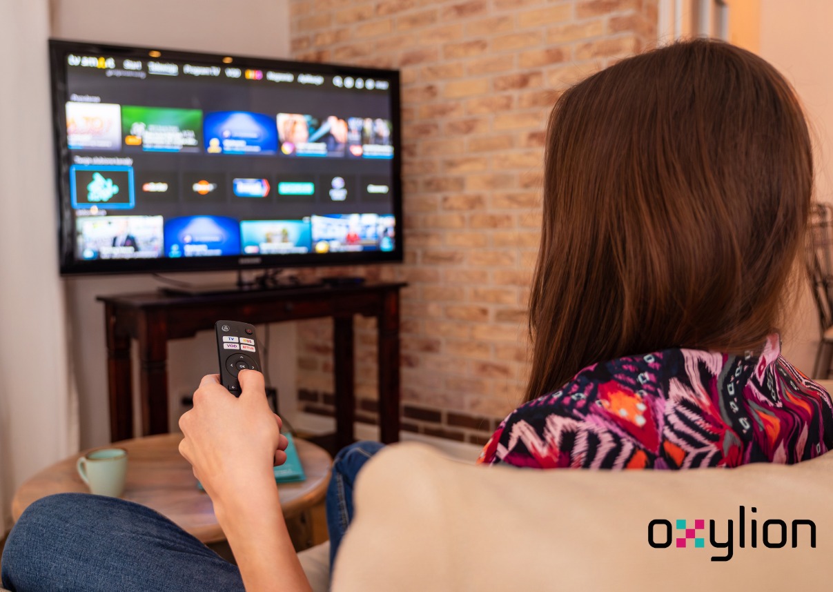 Kobieta z tyłu siedzi na kanapie, trzyma pilot i ogląda telewizor z interfejsem platformy streamingowej na tle ceglanej ściany.