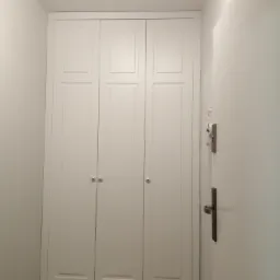 Biała, trzydrzwiowa szafa wnękowa z dekoracyjnymi gałkami, dopasowana do wysokości pomieszczenia w Luboniu. Na podłodze wzorzyste, szaro-beżowe płytki.