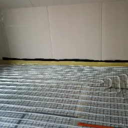 Białe rury ogrzewania podłogowego ułożone na czarnej siatce w pomieszczeniu z surowymi ścianami z płyt gipsowo-kartonowych. Widoczne żółte obrzeże i fragment pomarańczowych znaczników na rurach.