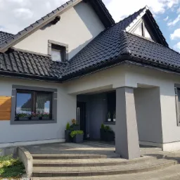 Nowoczesny dom jednorodzinny z szarą elewacją, czarnym dachem z dachówki ceramicznej i starannie zagospodarowanym terenem wokół wejścia, z widocznymi donicami z roślinami i kwiatami w oknach.