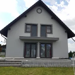 Szary dom jednorodzinny z ciemnobrązowym dachem i stolarką okienną, widok z poziomu trawnika, z elementami dekoracyjnymi z kamienia na schodach i obrzeżach.