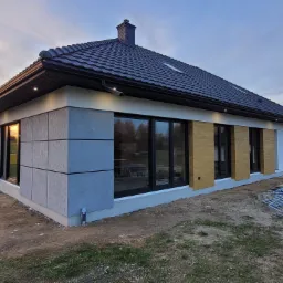 Nowoczesny dom jednorodzinny z elewacją wykończoną panelami w odcieniach szarości i drewna, czarną dachówką i oknami, oświetleniem zewnętrznym i rusztowaniem w tle.