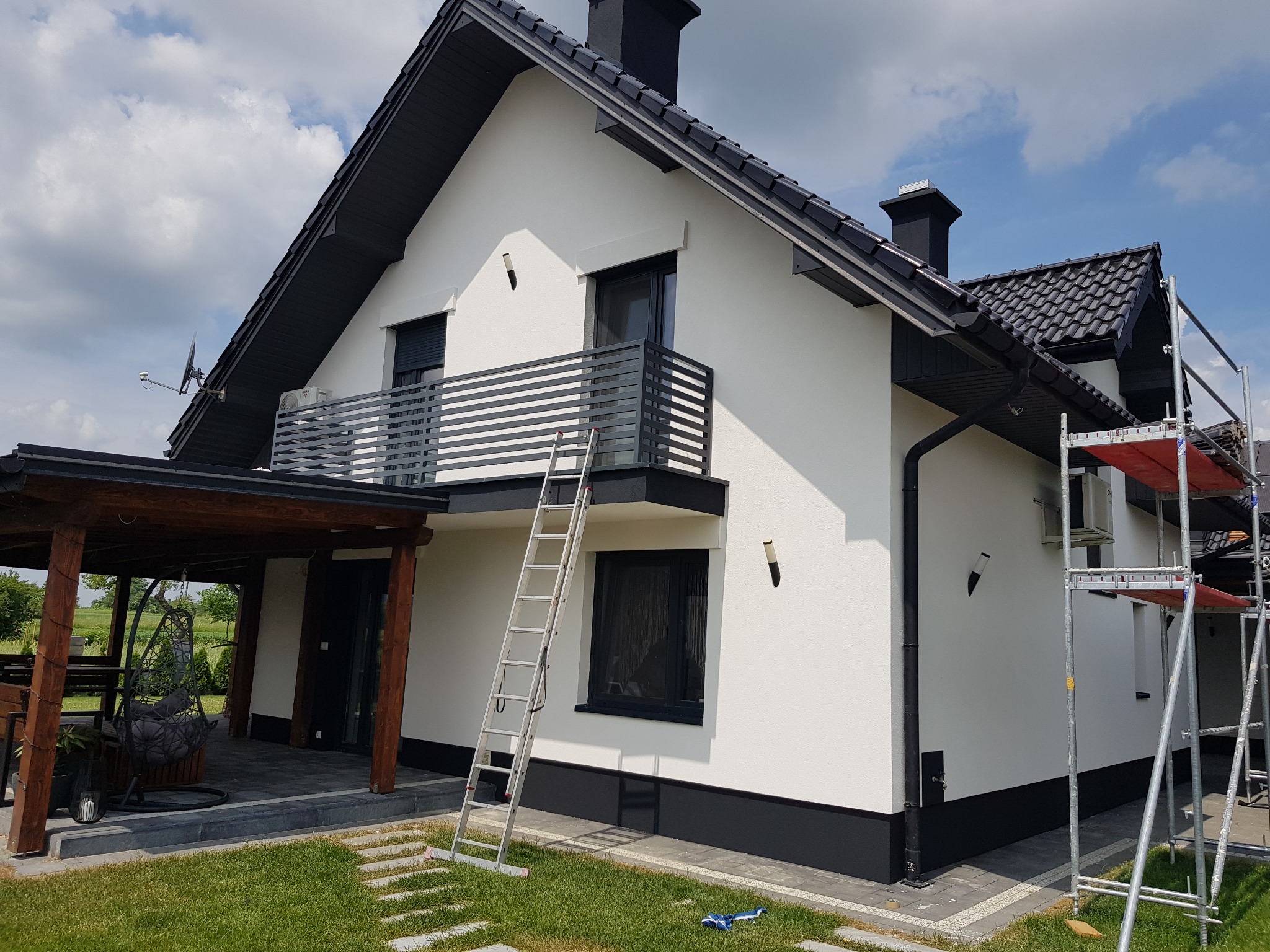 Dom jednorodzinny po malowaniu elewacji na biało z czarnymi elementami wykończeniowymi, widoczna drabina oparta o ścianę i rusztowanie z boku budynku, na trawniku leży niebieska szmatka.