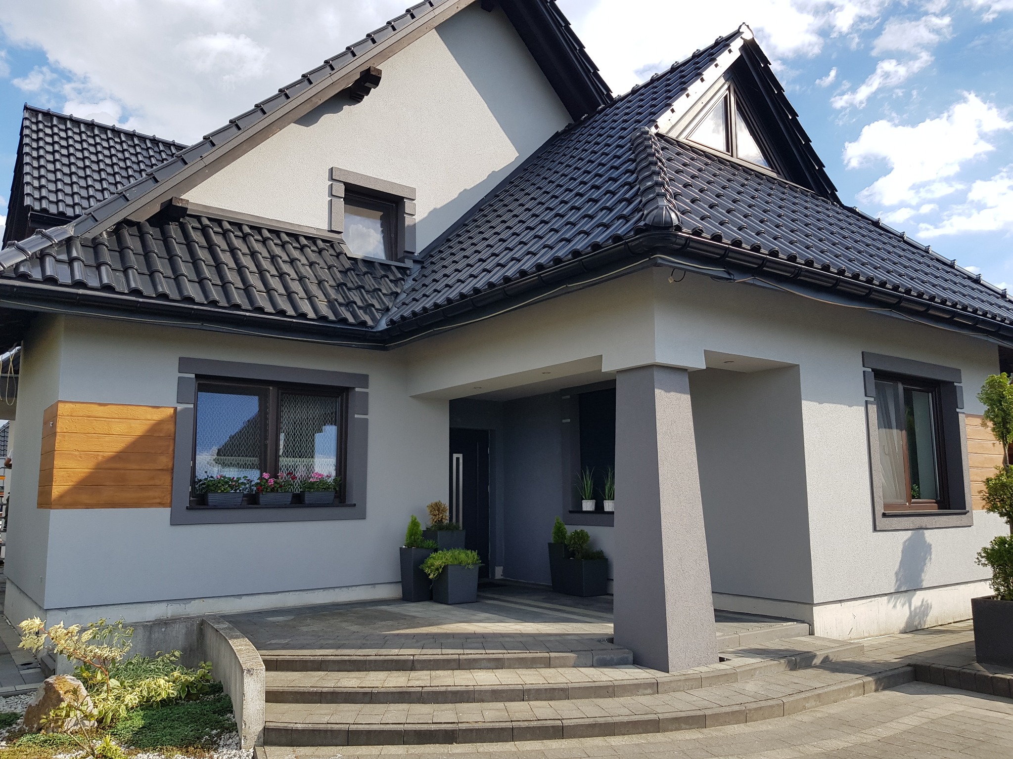 Nowoczesny dom jednorodzinny z szarą elewacją, czarnym dachem z dachówki ceramicznej i starannie zagospodarowanym terenem wokół wejścia, z widocznymi donicami z roślinami i kwiatami w oknach.