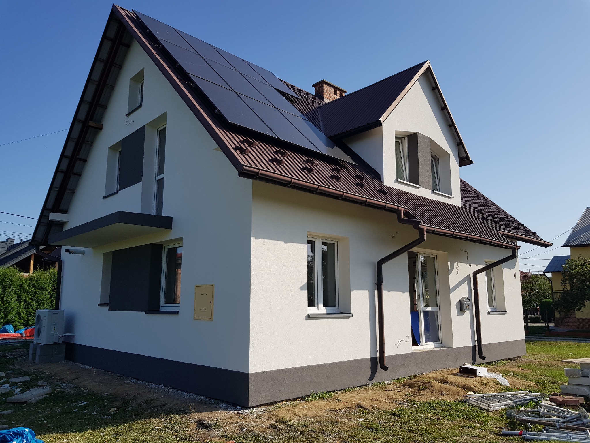 Nowoczesny dom jednorodzinny z jasną elewacją, ciemną podmurówką, brązowym dachem i panelami słonecznymi na dachu, uchwycony w słoneczny dzień.