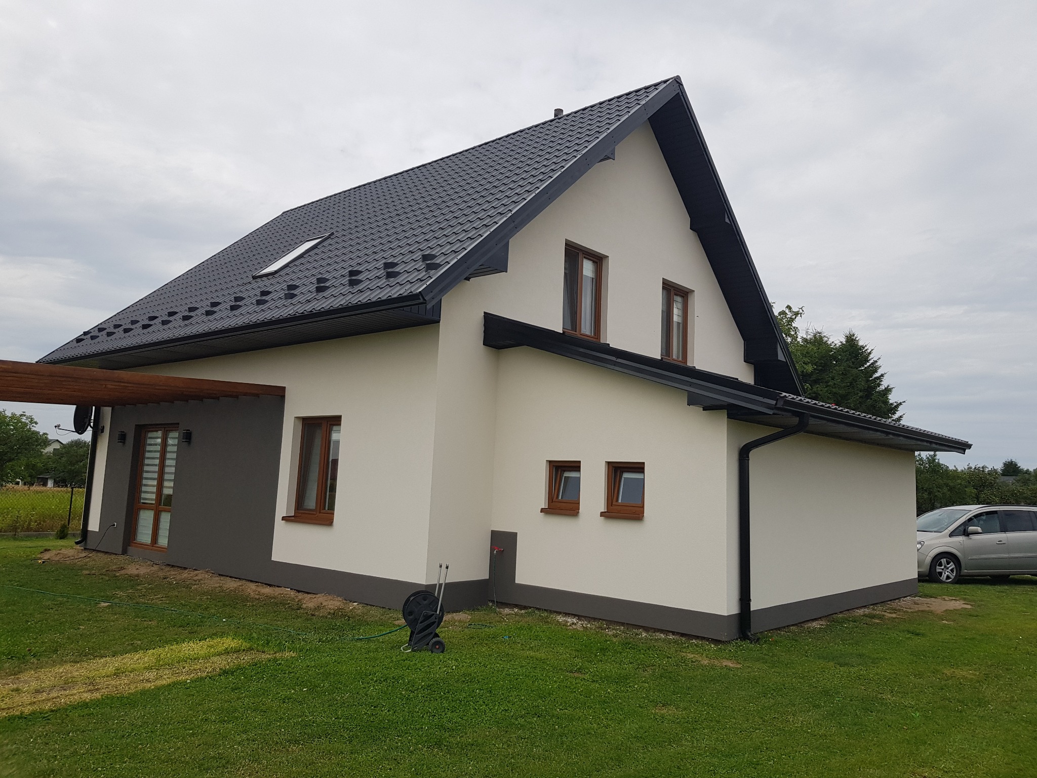 Nowoczesny dom jednorodzinny z jasną elewacją i ciemną podmurówką, czarnym dachem z lukarną, drewnianą pergolą i widoczną zwiniętą wężem ogrodowym na trawniku