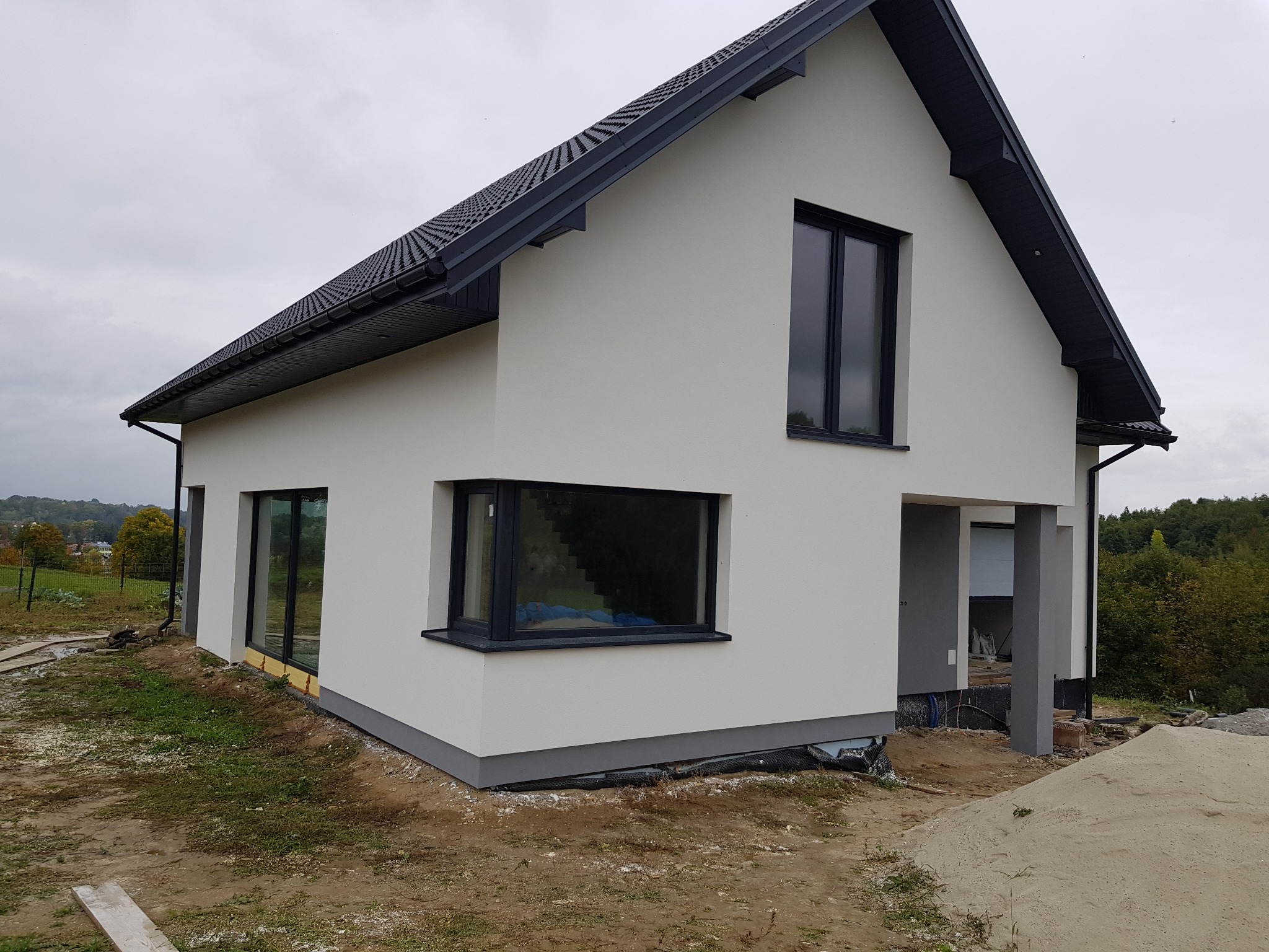 Nowoczesny dom jednorodzinny z jasną elewacją i ciemną stolarką okienną, widok z zewnątrz na etapie prac wykończeniowych terenu.