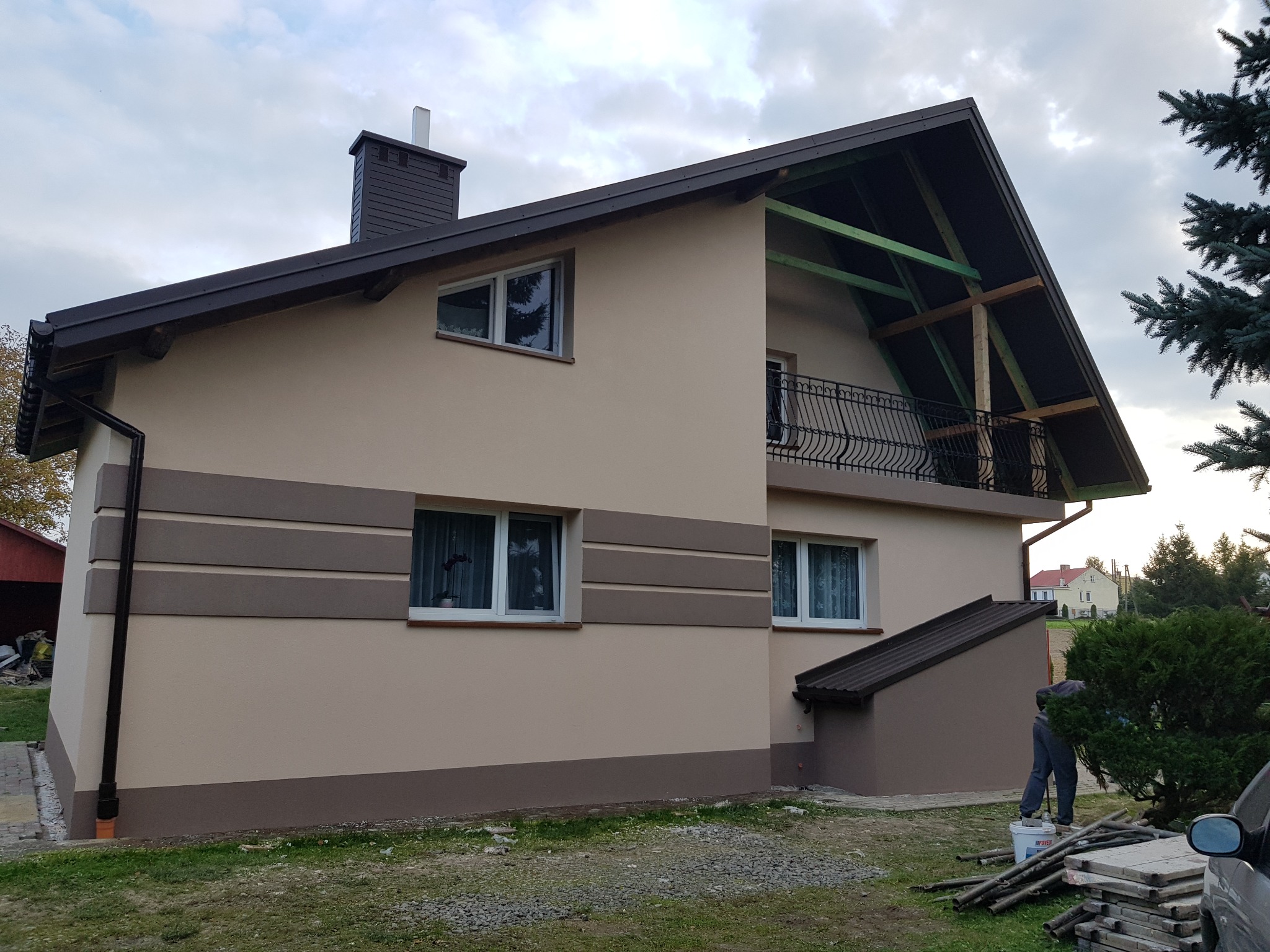 Dwukondygnacyjny dom po ociepleniu i malowaniu elewacji w odcieniach beżu i brązu, widoczny fragment niezakończonej elewacji pod dachem, pracownik podczas sprzątania na posesji.